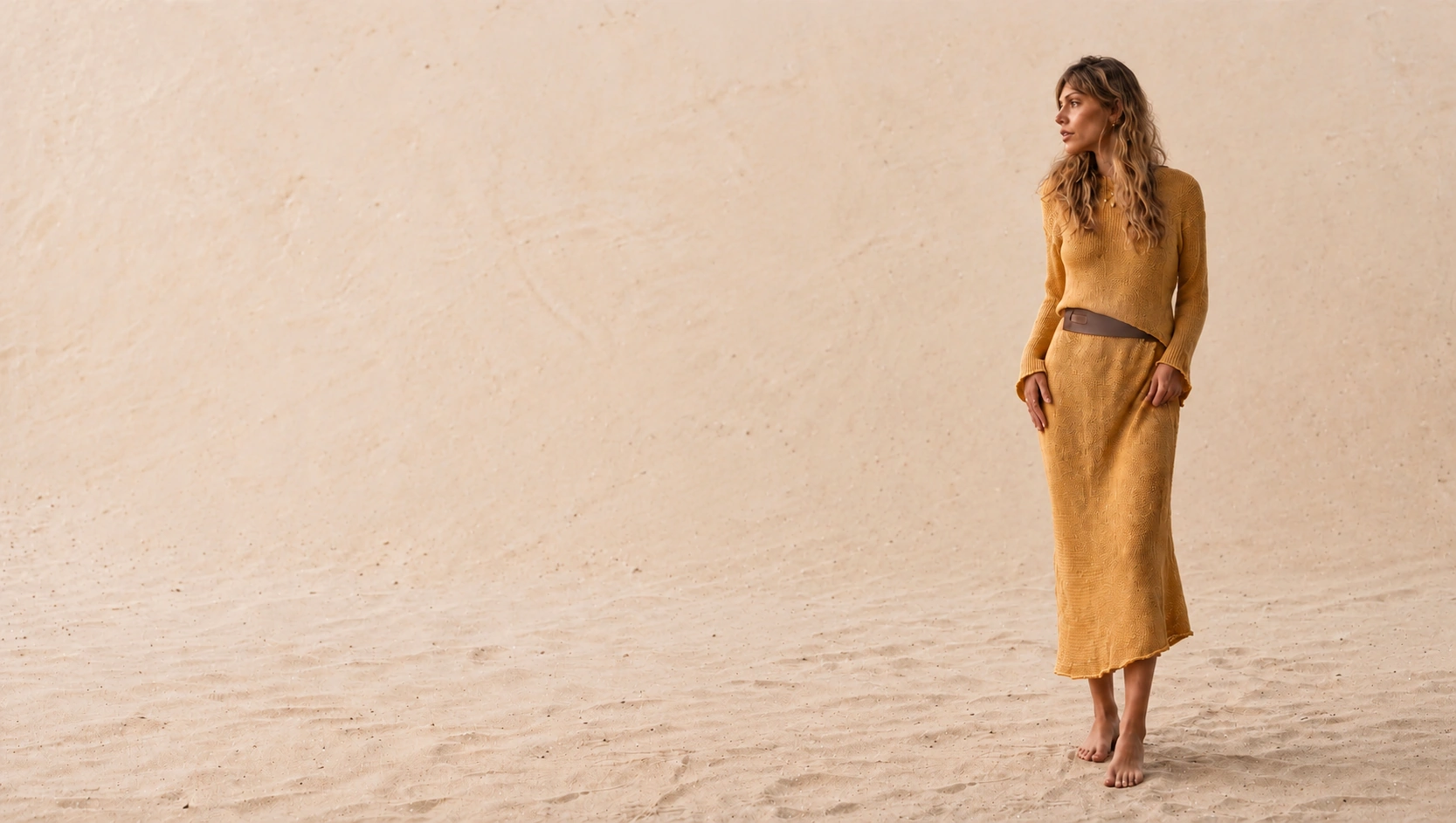 Modelo à direita, de corpo inteiro, com blusa e saia de tricô mostarda, cinto marrom e pés descalços, em cenário de areia e parede bege